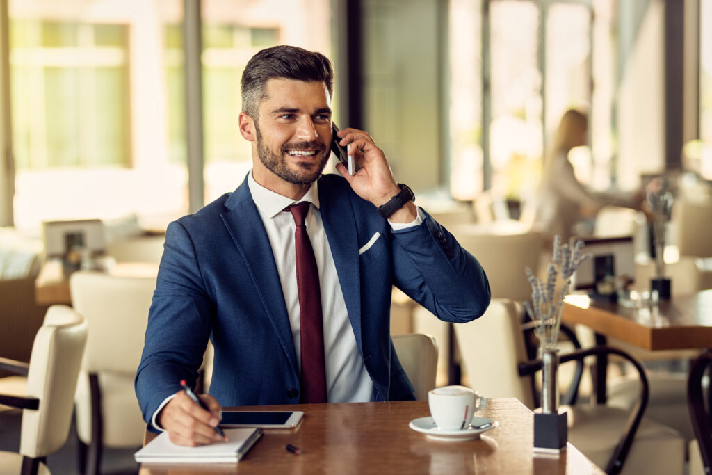 Happy businessman making a phone call and taking notes in a cafe.